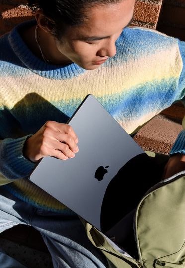 Một sinh viên đang ngồi bên ngoài. Anh ấy kéo một chiếc MacBook đóng nắp ra khỏi ba lô.